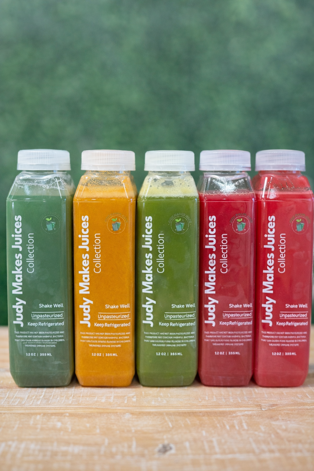 Five JMJ cold-pressed juice bottles in green, orange, and red on a wooden surface